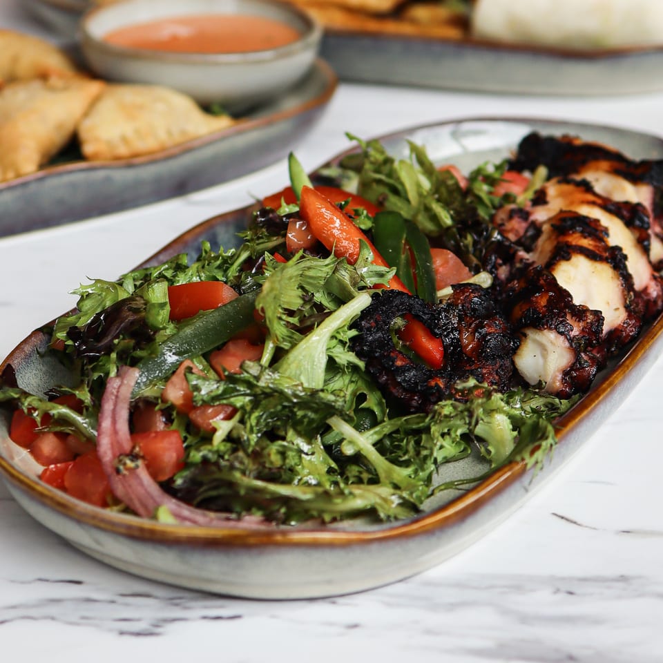 Grilled chicken and roasted vegetables served over a fresh mixed salad, with pastries and dipping sauce in the background.
