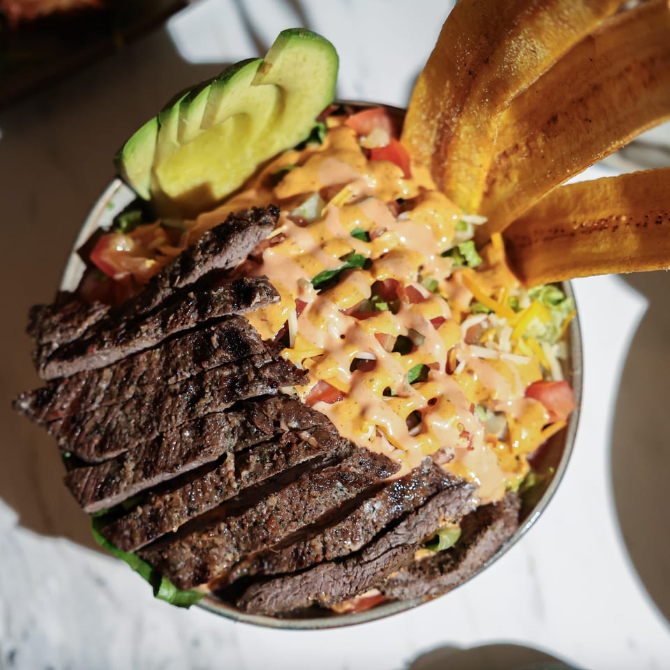Loaded salad bowl topped with sliced grilled steak, avocado, vegetables, creamy dressing, and crispy plantain chips.