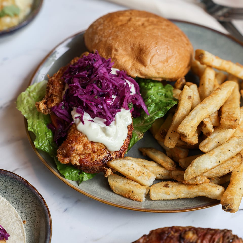 Crispy fried chicken sandwich with lettuce, purple cabbage slaw, and creamy sauce served alongside seasoned fries on a restaurant plate.