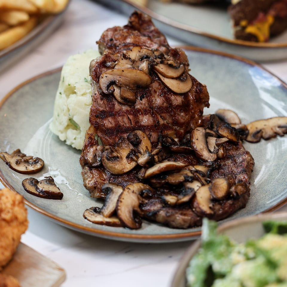 Grilled steaks topped with sautéed mushrooms and served with mashed potatoes on a blue plate, surrounded by side dishes.