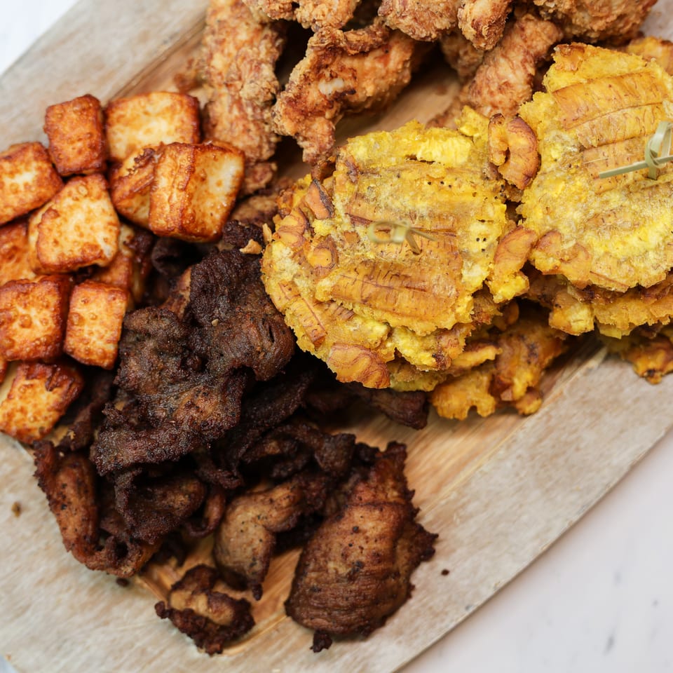 Wooden platter piled with fried chicken, golden plantains, crispy cheese cubes, and grilled meat.
